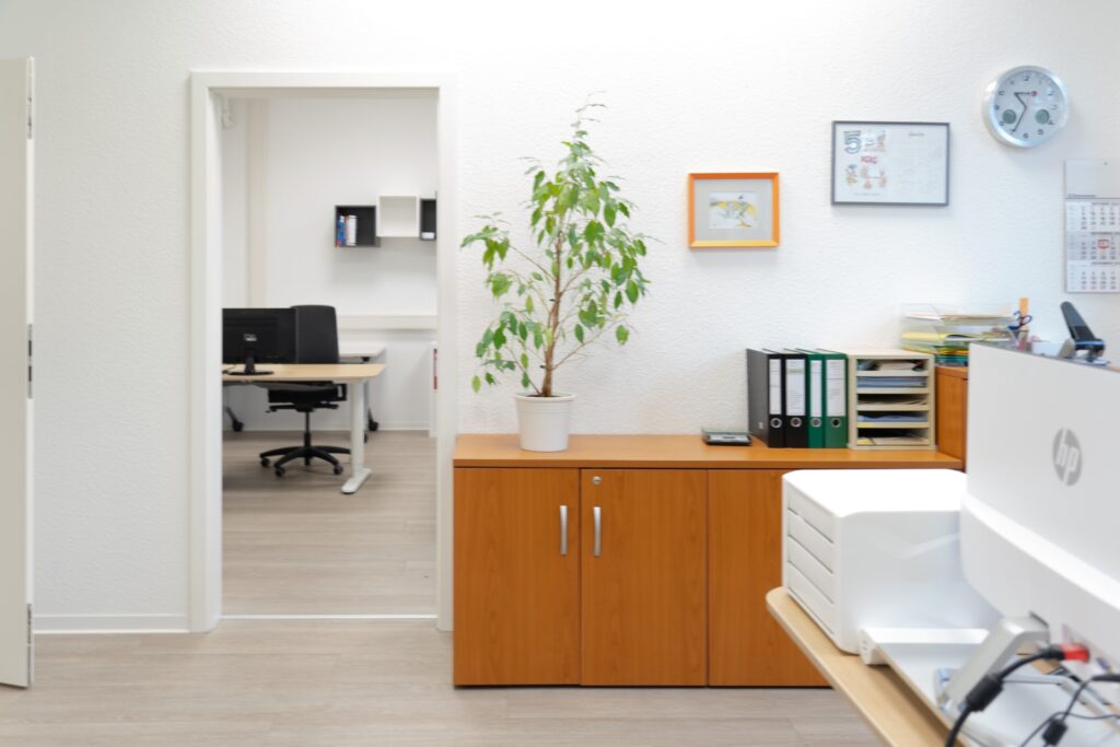 Helles Büro mit Schreibtisch, Aktenschränken, Zimmerpflanze und geöffnetem Besprechungsraum im Hintergrund