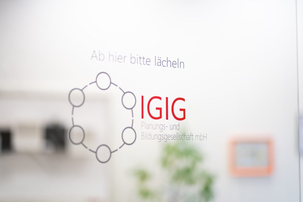 IGIG-Logo mit dem Spruch Ab hier bitte lächeln auf einer hellen Bürowand