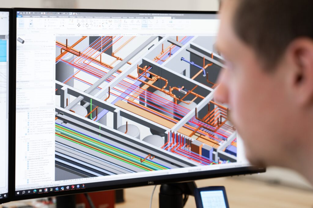 Mann betrachtet auf einem Monitor ein 3D-Gebäudemodell mit farbigen Leitungen und Rohren in Planungssoftware